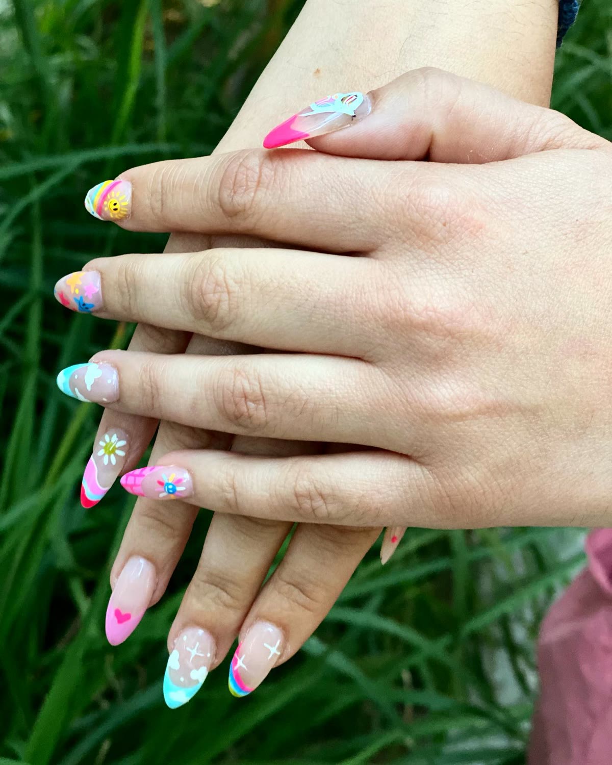 Two hands with pastel hand-painted floral nail art, photographed against greenery.